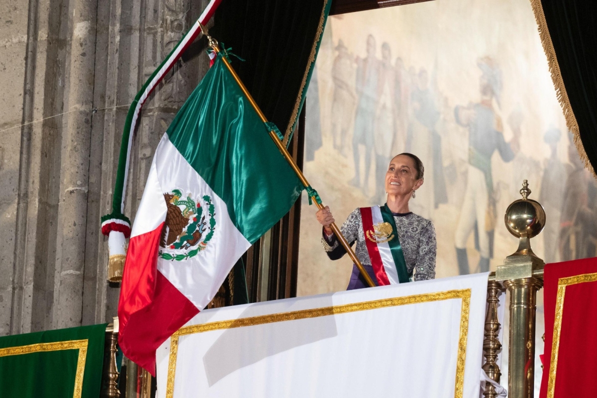 La presidenta ondea con orgullo la bandera de México desde el balcón principal de Palacio Nacional, enmarcada por la pintura histórica de los héroes de la Independencia.